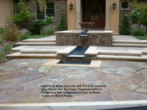 Light Sand Wash Concrete with Pre-Cast Concrete  Step Treads and Wall Caps. Flagstone Field in  Foreground with Ledgestone Veneer on Water  Feature to Match House 